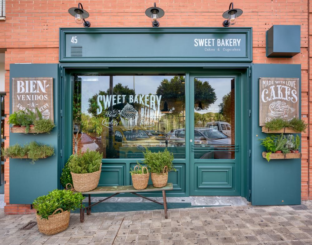 SWEET BAKERY, el obrador clásico inglés. En pleno barrio de Montequinto, siendo una antigua tienda de chucherías… Surge una imponente fachada verde carruaje completamente de madera y cristal, al más puro estilo ingles clásico. El diseño de un gran escaparate pintado a mano.Sweet Bakery es un pequeño obrador artesanal donde se elaboran grandes tartas, pasteles y cupcakes entre otros… Un diseño que destaca sobre la fachada de un edificio actual. Con puertas de hierro que se abren y adornadas con plantas y cestos de mimbre.Todo un concepto natural, artesanal y clásico con paredes de ladrillo antiguo, moldurones, zócalos. Un suelo hidráulico gastado con bancos y tapas de madera vieja, nos transporta 100 años atrás. Incluyendo una carpintería que nos permite ver el trabajo que se realiza de obrador en su interior.